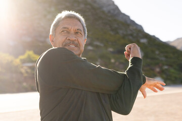 Happy senior biracial man wearing sportswear, stretching on street in mountains