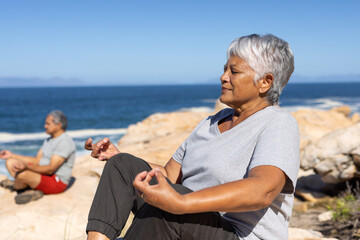 Happy senior biracial couple sitting on rocks by the sea, doing yoga and meditating