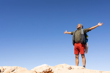 Happy senior biracial man hiking, widening arms in mountains, with copy space