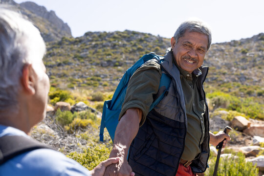 Happy senior biracial couple wearing backpacks, holding hands and hiking with trekking poles