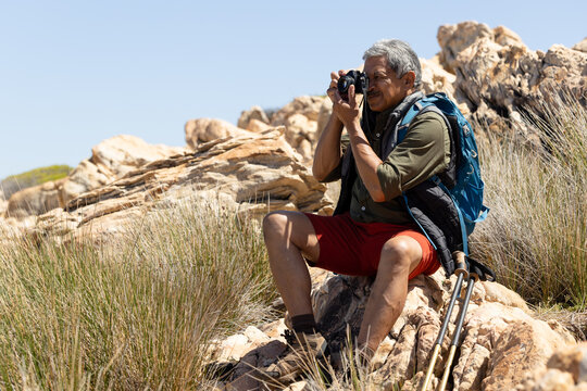 Happy senior biracial man wearing backpack, hiking in mountains, taking pictures with camera