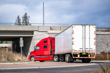 Bonnet red big rig semi truck transporting cargo in dry van semi trailer turning on the round road intersection going under the bridge