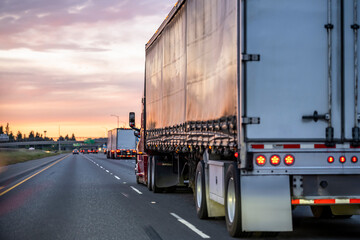 Burgundy big rig semi truck with black covered semi trailer driving on the highway road at twilight...