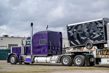 Fototapeta na wymiar Violet classic big rig American bonnet semi truck with loaded step down semi trailer standing on the industrial warehouse parking lot waiting for the next freight start
