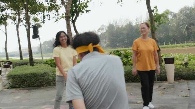 Handheld camera shot of blindfolded Asian man smiling and trying to catch family members while playing blind mans buff game together in park