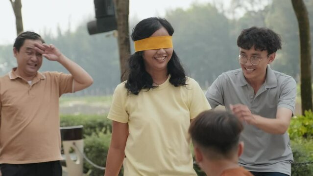 Handheld camera shot of blindfolded Asian woman smiling and playing blind mans buff game with family in park, catching her son, laughing and hugging him