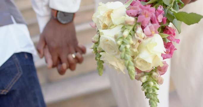 Midsection of african american gay male couple holding hands and flowers at wedding, slow motion