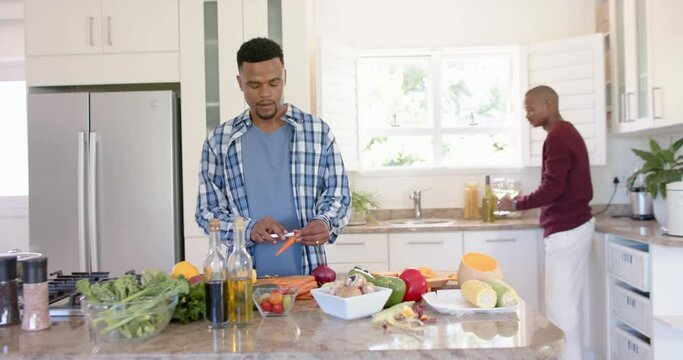Happy african american gay male couple preparing dinner, drinking wine in kitchen, slow motion