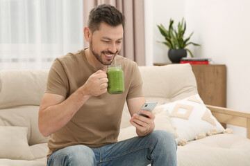 Happy man holding delicious fresh smoothie and using smartphone at home - Powered by Adobe