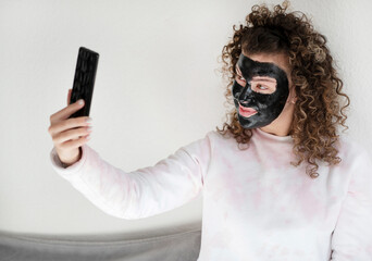 Young beautiful woman with acne problem skin, applies dark cosmetic mask and makes selfie on her mobile phone