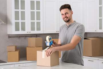 Smiling man taping box with adhesive tape dispenser in kitchen, space for text