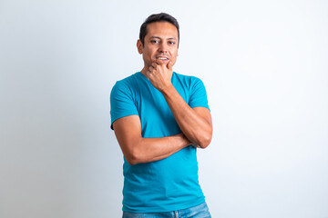 Hombre joven moreno, con ropa casual playera azul, sonriendo y tocando la barbilla con fondo blanco