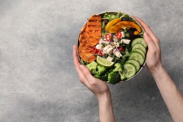 Woman holding bowl with many different vegetables and tofu at grey table, top view and space for text. Vegan diet