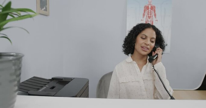 Portrait of medical receptionist sitting at front desk, talking on telephone, slow motion