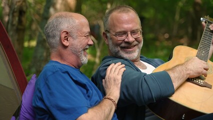 Closeup of two gay men enjoying nature camping with tent with pride flag in forest on river playing guitar.