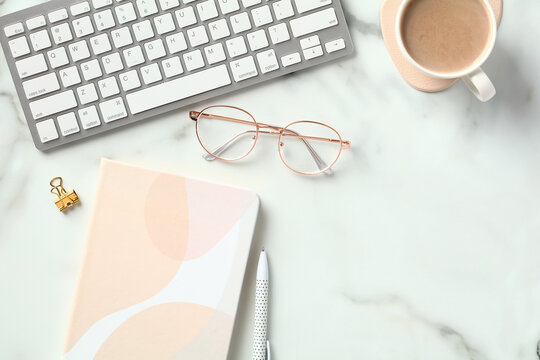 Modern office desk table top view. Flat lay keyboard, cup of coffee, paper notebook, glasses on marble background.