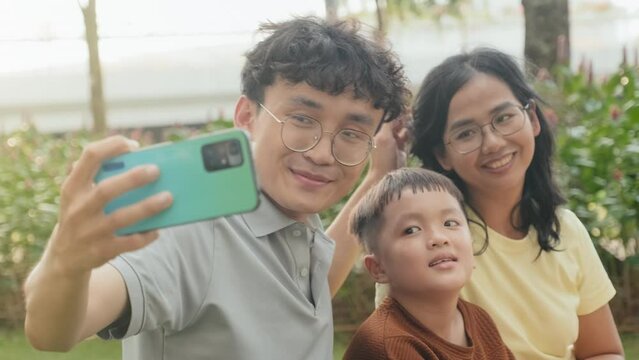 Rack focus shot of happy Asian parents and little son smiling and posing for smartphone camera while taking selfie on summer day in park