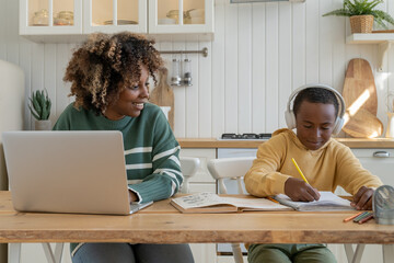 Smiling young African American mother doing homework with small son, sitting at kitchen table together. Black woman mom helping child with home task assignment while working remotely on laptop