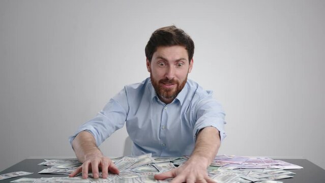 A man in a blue shirt piling up a bunch of money and laying ontop of it. He looks really happy. The man is in light gray room. High quality 4k footage