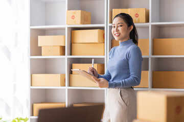 Asian Female entrepreneur starting a small business for SME online sales working with parcel boxes at home checking orders from the internet preparing for SME delivery.