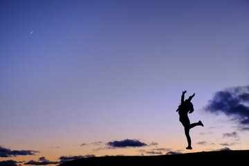 A silhouette of a woman in gym uniform jumping on top of a hill in celebration after completing her exercise with blue and orange sky in the background with a crescent moon at the top left corner.