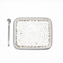 On a white background, there is a silver tray with round, oval tablet pills and a silver metal spoon next to it.