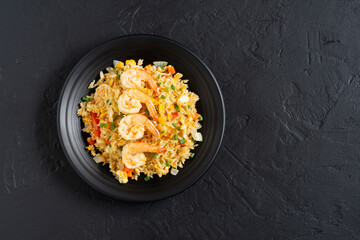 Fried rice with vegetables and sea prawns, carrots, eggs, and tomatoes. Thai Chinese food is arranged on a black plate on a black background kitchen table, top view.