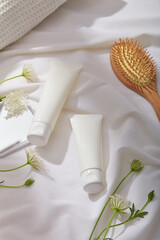 Two empty label tubes displayed on white fabric background. A wooden brush, hand mirror and some white flowers. mockup of skin care cosmetic tube of beauty facial