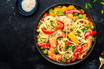 Stir fry egg noodles with shrimps, paprika, green pea, chives and sesame seeds bowl. Asian cuisine dish. Black kitchen table background, top view