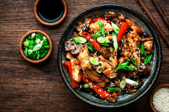 Stir fry chicken with paprika, mushrooms, green chives and sesame seeds in ceramic bowl.  Asian cuisine dish. Wooden kitchen table background, top view