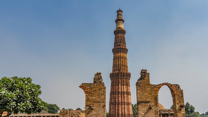Fototapeta na wymiar The world's tallest brick minaret against the blue sky. Nearby - dilapidated walls, arched openings. Ruins of the ancient temple complex Qutub Minar. India. Delhi