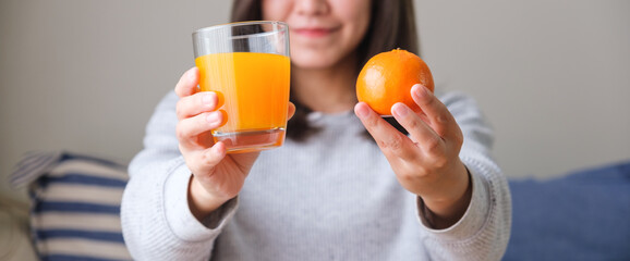 Closeup image of a young woman holding and showing an orange and a glass of fresh orange juice at home