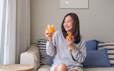 Fototapeta premium Portrait image of a young woman holding an orange and a glass of fresh orange juice at home