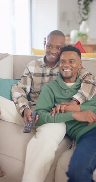 Vertical video of african american gay male couple sitting on sofa and watching tv, slow motion