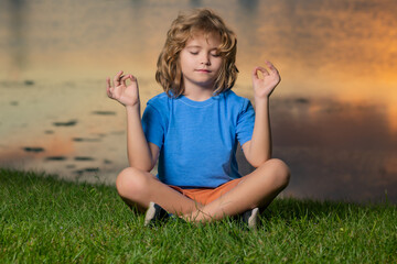 Fototapeta premium Child meditating in lotus positionon on grass, relaxing on summer nature. Happy little kid meditates on green grass at summer day.