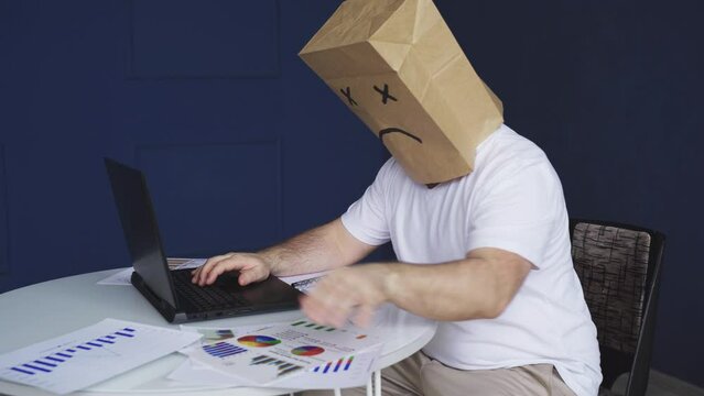 A male businessman in a white shirt with a paper bag on his head, with a sad smiley face drawn, works at a computer, with documents. Emotions and gestures.