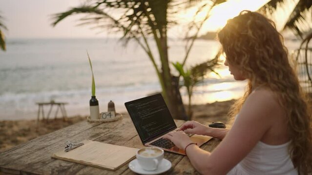 Young female developer working on laptop by the ocean. Woman freelancer coding at outdoor tropical cafe at sunset. Caucasian girl working remotely typing on computer at exotic location. Worldwide work