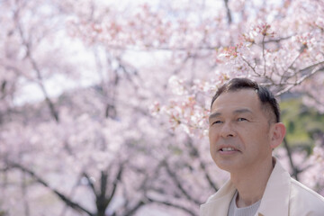 Middle-aged Asian man wearing white cardigan with cherry blossoms in background.