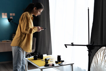 Female product photographer with smartphone and camera photographing cosmetic products in home photo studio. Professional and mobile photo
