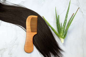 Hair care wooden comb on a long strand of black hair with Aloe Vera hair care on background. Tools...
