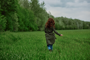 a woman in a long raincoat runs across a field in tall green grass in cloudy weather in spring