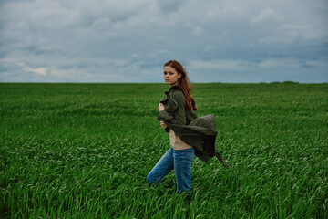 Naklejka na ściany i meble a red-haired woman stands in a green field in rainy, cold weather, holding a raincoat in the wind
