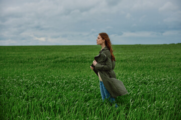 Fototapeta premium a red-haired woman stands in a green field in rainy, cold weather, holding a raincoat in the wind