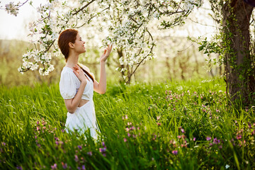 a beautiful woman in a light dress stands next to a flowering tree and smells the flowers