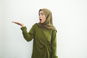 Smiling beautiful asian woman in green dress and hijab pointing to copy space with palms, showing advertising products and confidently looking at camera isolated over white background.