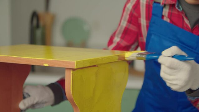 Close-up of black DIY woman in protective workwear and gloves restoring and renewing old furniture for reuse , painting stool with paintbrush in yellow color indoors.