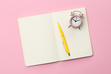 Ballpoint pen, notebook and alarm clock on pink background, top view