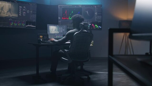 Handheld shot of African American colorist doing color grading on computer in modern studio. Multiple big monitors with action movie footage and program interface on the wall. Video post production.