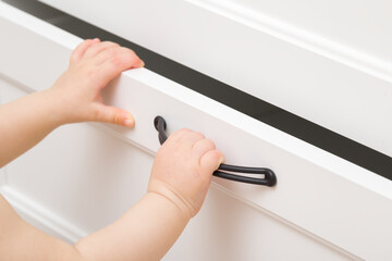 Baby hands open or close white drawer at home room. Closeup. Side view. Unsafe using wooden furniture.