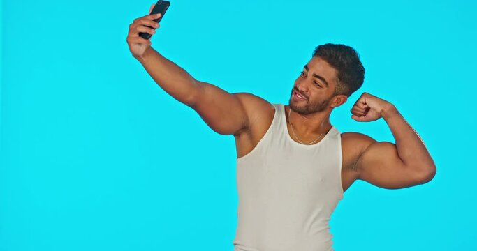 Selfie, smile and bicep muscle of man in studio isolated on a blue background for fitness. Photographer, flexing emoji and happy male model taking photo for profile picture, social media and workout.